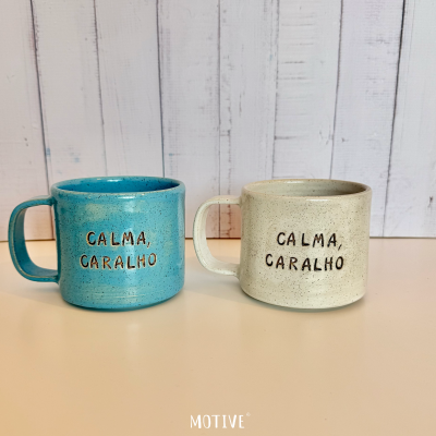 Canecas de cerâmica azul turquesa e bege com texto CALMA, CARALHO em fundo branco de madeira.