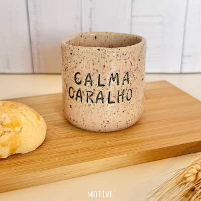 Copo de cerâmica bege manchado com frase CALMA CARALHO, sobre tábua de madeira com pão e espigas de trigo.