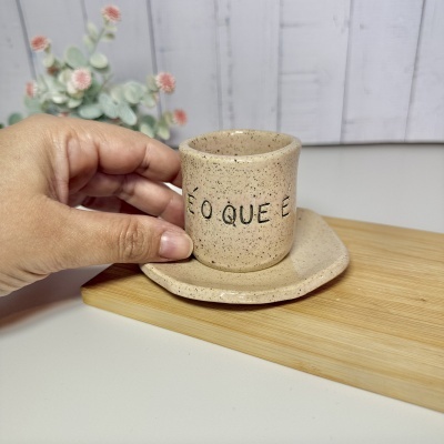 Conjunto de chávena e prato em cerâmica bege com escrita, mão a segurar chávena sobre superfície de madeira.