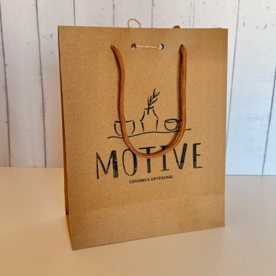 Saco de papel castanho com alças de corda laranja e texto MOTIVE CERÂMICA ARTESANAL
