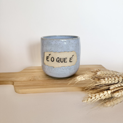 Copo cerâmico azul com etiqueta É O QUE É sobre tábua de madeira clara e trigo seco ao lado