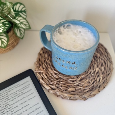 Caneca azul com texto CALMA, CARALHO e espuma de leite sobre base de palha com leitor digital e planta
