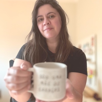 Caneca de cerâmica branca com texto, segurada por mulher em ambiente interior.