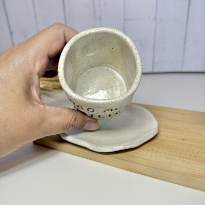 Caneca cerâmica bege com pontos castanhos e base larga segurada por uma mão