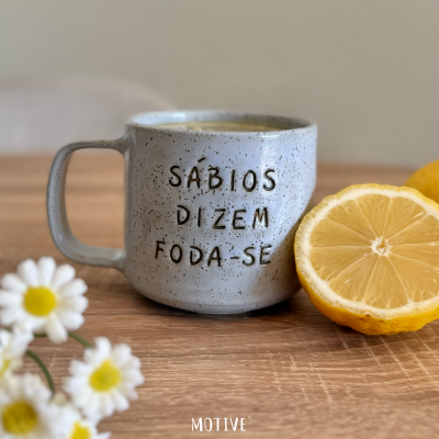 Caneca de cerâmica cinzenta com frase gravada e meio limão ao lado numa mesa de madeira clara.