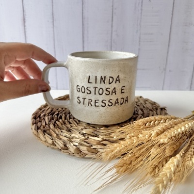 Caneca de cerâmica bege com frase gravada e base de fibras naturais com trigo ao lado