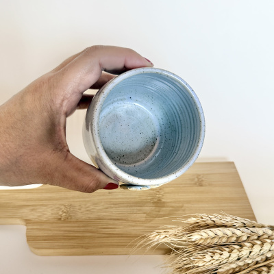 Vaso cerâmico azul segurado por mão com unhas vermelhas, sobre mesa de madeira clara, perto de espigas de trigo secas