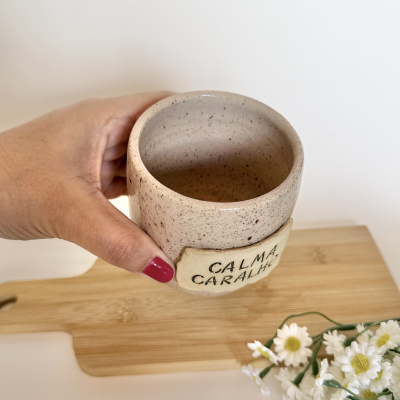 Caneca de cerâmica bege com manchas castanhas e etiqueta CALMA CARALHO