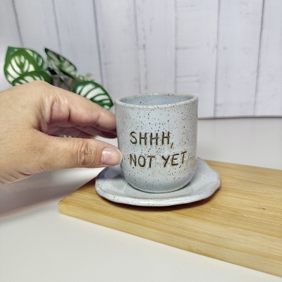 Caneca cinza com texto SHHH, NOT YET em cerâmica pontilhada com pires, mão segurando, fundo branco e planta