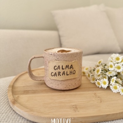 Caneca de cerâmica rosa com texto CALMA, CARALHO numa bandeja de madeira com flores brancas e amarelas ao lado