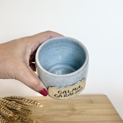 Caneca de cerâmica azul com placa bege escrita CALMA CALHÃO segura por mão com unha vermelha