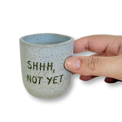 Caneca cerâmica cinzenta com texto SHHH, NOT YET segurada por mão