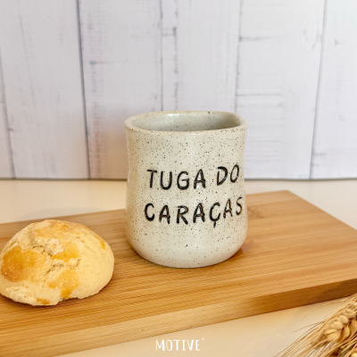 Caneca de cerâmica bege com texto TUGA DO CARAÇAS em relevo numa tábua de madeira com pão e espigas secas