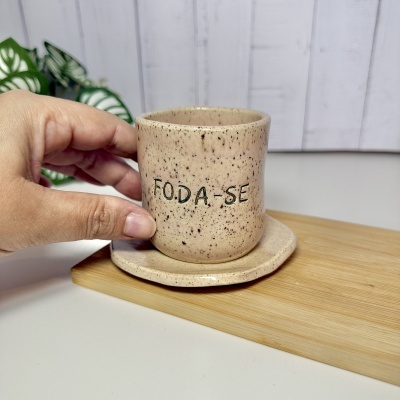Caneca de cerâmica bege com o texto FODA-SE e pires sobre base de madeira.