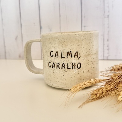 Caneca cerâmica bege com texto CALMA, CARALHO e espigas de trigo secas