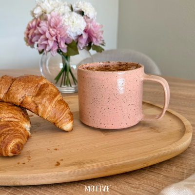 Caneca rosa em tabuleiro de madeira com croissants e vaso com flores em fundo neutro.