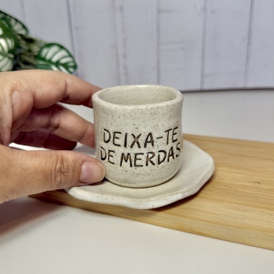 Caneca pequena em cerâmica branca com pontos castanhos e frase gravada, segurada por mão, sobre base branca e tábua de madeira clara