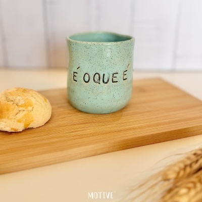 Caneca de cerâmica verde com texto É O QUE É sobre tábua de madeira com pão ao lado.