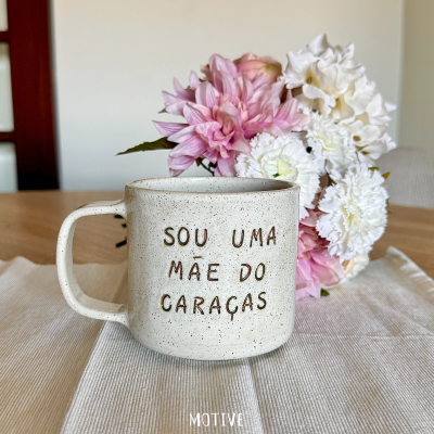 Caneca cerâmica bege com texto gravado e flores rosas e brancas ao fundo