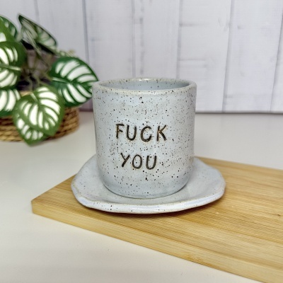 Caneca em cerâmica cinzenta com texto e prato sobre base de madeira com planta atrás