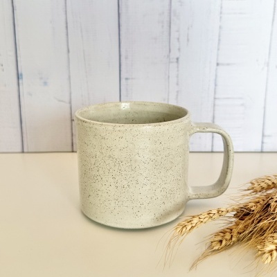 Caneca bege claro com manchas sobre superfície clara e espigas de trigo ao lado
