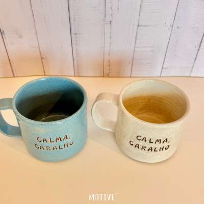 Duas canecas de cerâmica com texto CALMA, CARALHO, uma azul e outra bege, sobre superfície clara com fundo de madeira branca.