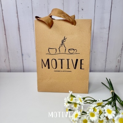 Saco de papel com alças castanho com texto MOTIVE CERÂMICA ARTESANAL e flores brancas e amarelas ao lado