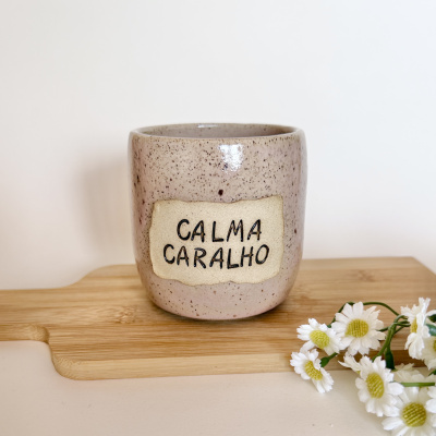 Caneca de cerâmica bege com etiqueta CALMA CARALHO perto de flores brancas e base de madeira.