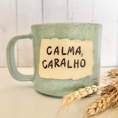 Caneca verde cerâmica com placa bege e texto CALMA, CARALHO ao lado de espigas de trigo