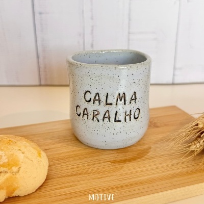 Caneca de cerâmica cinza com texto CALMA CARALHO sobre tábua de madeira com pão e espigas de trigo