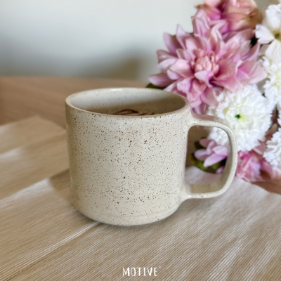 Caneca de cerâmica bege com textura manchada e flores cor-de-rosa ao fundo