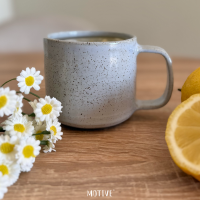 Caneca de cerâmica cinzenta com manchas pretas sobre mesa de madeira com flores e limão