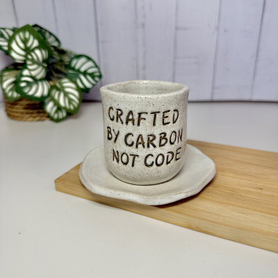 Caneca cerâmica branca com texto CRAFTED BY CARBON NOT CODE e planta verde ao fundo
