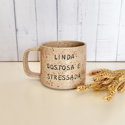 Caneca de cerâmica bege com texto LINDA GOSTOSA E STRESSADA ao lado de espigas de trigo