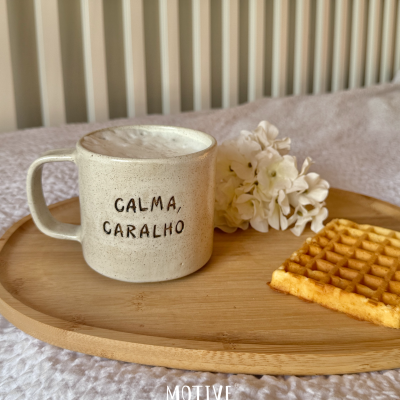 Caneca de cerâmica bege com frase CALMA, CARALHO em tabuleiro de madeira com waffle e flores brancas