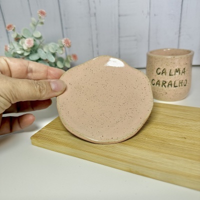 Pequeno prato e caneca de cerâmica bege com frase CALMA CARALHO em relevo, com flores ao fundo.