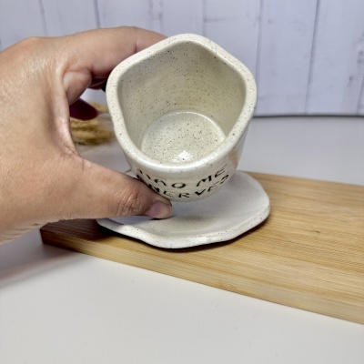 Copo de cerâmica hexagonal creme com pintas castanhas e pires na mão sobre tábua de madeira.