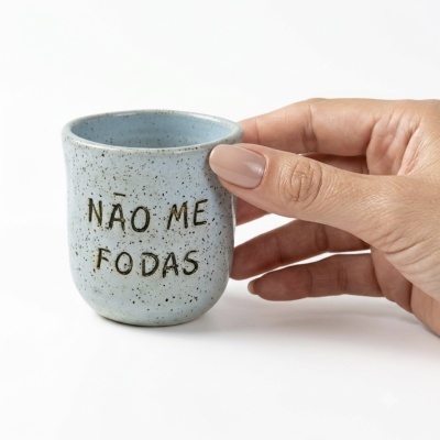 Mini caneca azul claro em cerâmica com texto NÃO ME FODAS, segurada por mão com unhas nude.