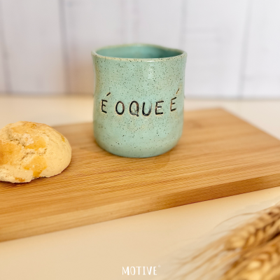Caneca cerâmica verde com texto É O QUE É ao lado de pão e espigas de trigo sobre tábua de madeira