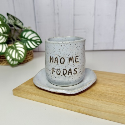 Copo cerâmico cinza claro com texto NÃO ME FODAS sobre pires cinza, sobre tábua de madeira, com planta ao lado.