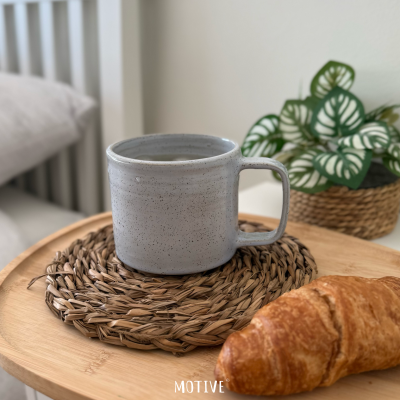 Caneca cerâmica cinzenta sobre base de palha com croissant e planta em vaso trançado
