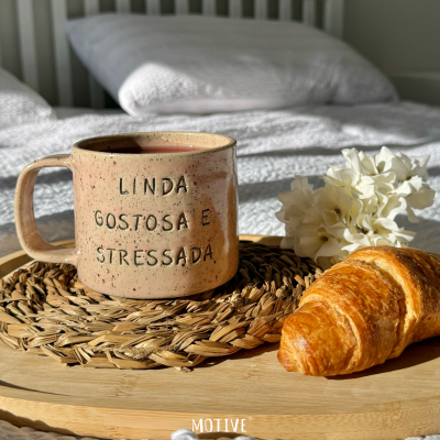 Caneca de cerâmica bege com texto sobre base de madeira com croissant e flores, em cama branca