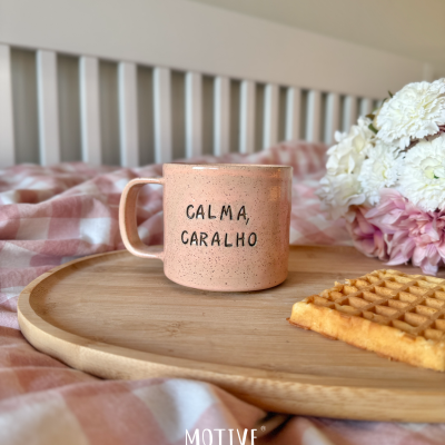 Caneca rosa claro com texto CALMA, CARALHO numa tábua de madeira, junto com uma bolacha waffle e flores, sobre cama com padrão xadrez rosa e branco.