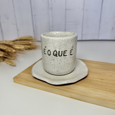 Caneca de cerâmica branca com manchas pretas e texto em português sobre pires em madeira