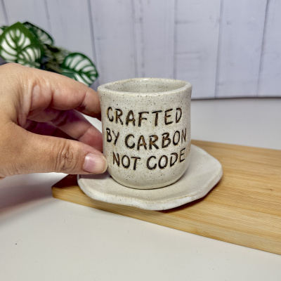 Caneca de cerâmica branca com texto em relevo segura por uma mão sobre pires branco em base de madeira clara.