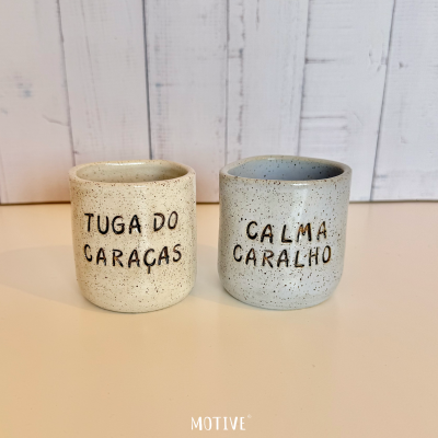 Duas canecas de cerâmica salpicada com textos em preto sobre fundo branco de madeira.
