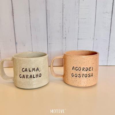Duas canecas de cerâmica com frases em português, uma bege e outra rosa, com fundo de madeira pintada de branco.