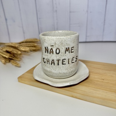 Caneca de cerâmica bege com texto preto NÃO ME CHATEIES sobre prato e tábua de madeira clara.