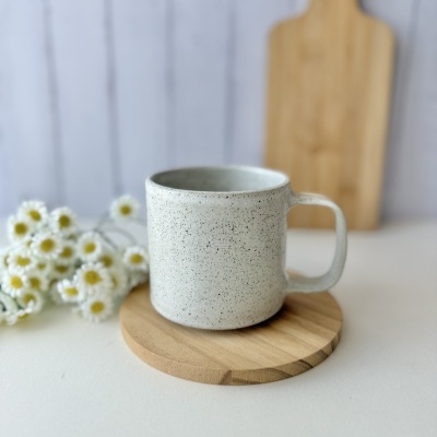 Caneca de cerâmica branca salpicada com pega quadrada sobre base de madeira com flores ao lado.