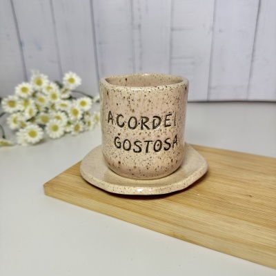 Caneca de cerâmica bege com texto sobre pires, tábua de madeira e flores brancas ao fundo